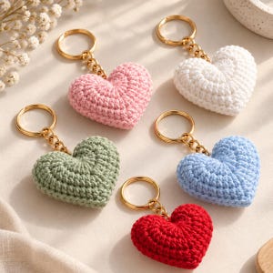 May include: Five heart-shaped keyrings made of crocheted yarn in pastel pink, white, sage green, light blue, and bright red. Each heart is attached to a gold-toned metal split ring by a short chain, creating a collection of five distinct items.