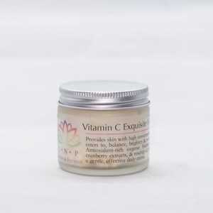 May include: A white jar with a silver lid labeled "Vitamin C Exquisite Face Cream". The label also includes a pink and purple lotus flower design and the text "Provides skin with high concentrations of Vitamin C esters to, balance, brighten & revitalize. Antioxidant-rich organic blueberry, cranberry extracts, & rosehip oil create a gentle, effective daily cr&egrave;me." The text "NP" and "ONS & POTIONS" are also visible on the label.