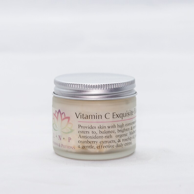 May include: A white jar with a silver lid labeled "Vitamin C Exquisite Face Cream". The label also includes a pink and purple lotus flower design and the text "Provides skin with high concentrations of Vitamin C esters to, balance, brighten & revitalize. Antioxidant-rich organic blueberry, cranberry extracts, & rosehip oil create a gentle, effective daily cr&egrave;me." The text "NP" and "ONS & POTIONS" are also visible on the label.