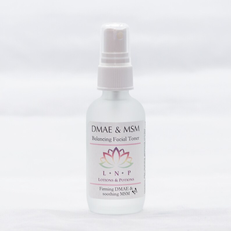 May include: A clear glass bottle with a white spray top and label. The label reads "DMAE & MSM Balancing Facial Toner" with a pink and purple lotus flower graphic. Below the flower is the text "LNP LOTIONS & POTIONS" and "Firming DMAE & soothing MSM".