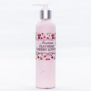 May include: A pink bottle of Montana Flathead Cherry Lotion with a silver pump top and a label featuring red cherries and green leaves. The label reads "Montana Flathead Cherry Lotion" and "All Natural".