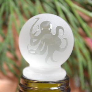 May include: A frosted glass bottle stopper with an etched design of an octopus. The stopper is white and the octopus is a dark gray.