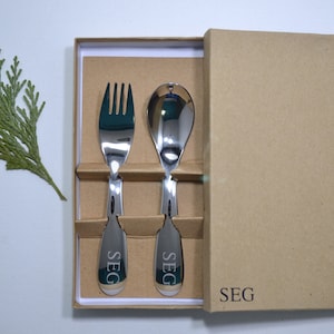 May include: A set of stainless steel fork and spoon in a brown cardboard box. The utensils have the word "SEG" engraved on the handles.