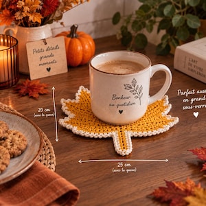 Puede incluir: Un posavasos de ganchillo color amarillo mostaza con forma de hoja de arce y borde color crema. El posavasos mide 20 cm de largo sin el tallo y 25 cm de ancho con el tallo. Una taza de cerámica con el texto Bonheur au quotidien está encima.