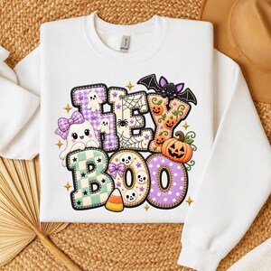 May include: A white long-sleeve sweatshirt featuring a colorful graphic with the text HEY BOO. The design includes a ghost with a purple bow, a bat, pumpkins, skulls, a spider web, and candy corn, all rendered in a playful, stitched-style pattern.