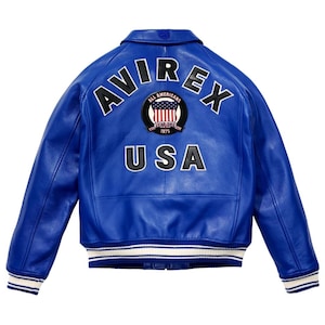 Op de afbeelding: Een felblauw leren jack met een omgeslagen kraag en lange mouwen. Op de achterkant staat de tekst AVIREX in zwarte letters, een ronde patch met een Amerikaanse vlag en daaronder de tekst USA. De zoom en boorden hebben een gestreept patroon.