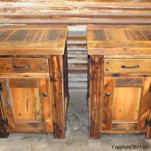 May include: Two rustic wooden nightstands with a drawer and a door. Each nightstand has a distressed wood finish and black metal hardware. The nightstands are made from reclaimed wood.