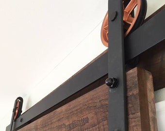 May include: Close-up of a black metal sliding barn door track with copper-colored wheels. The track is mounted to a wooden door and a white ceiling.