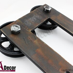 May include: A U-shaped metal bracket with a rusted finish, featuring two black spoked wheels attached by silver hexagonal bolts. The metal surface is stamped with the number 1776. The bracket has a flat, rectangular frame design.