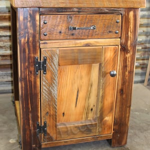 May include: A rustic wooden cabinet with a single drawer and a door. The cabinet is made of reclaimed wood and has a dark brown finish. The door has a simple design with a single panel and a black metal handle. The drawer has a black metal pull. The cabinet is sitting on a concrete floor.