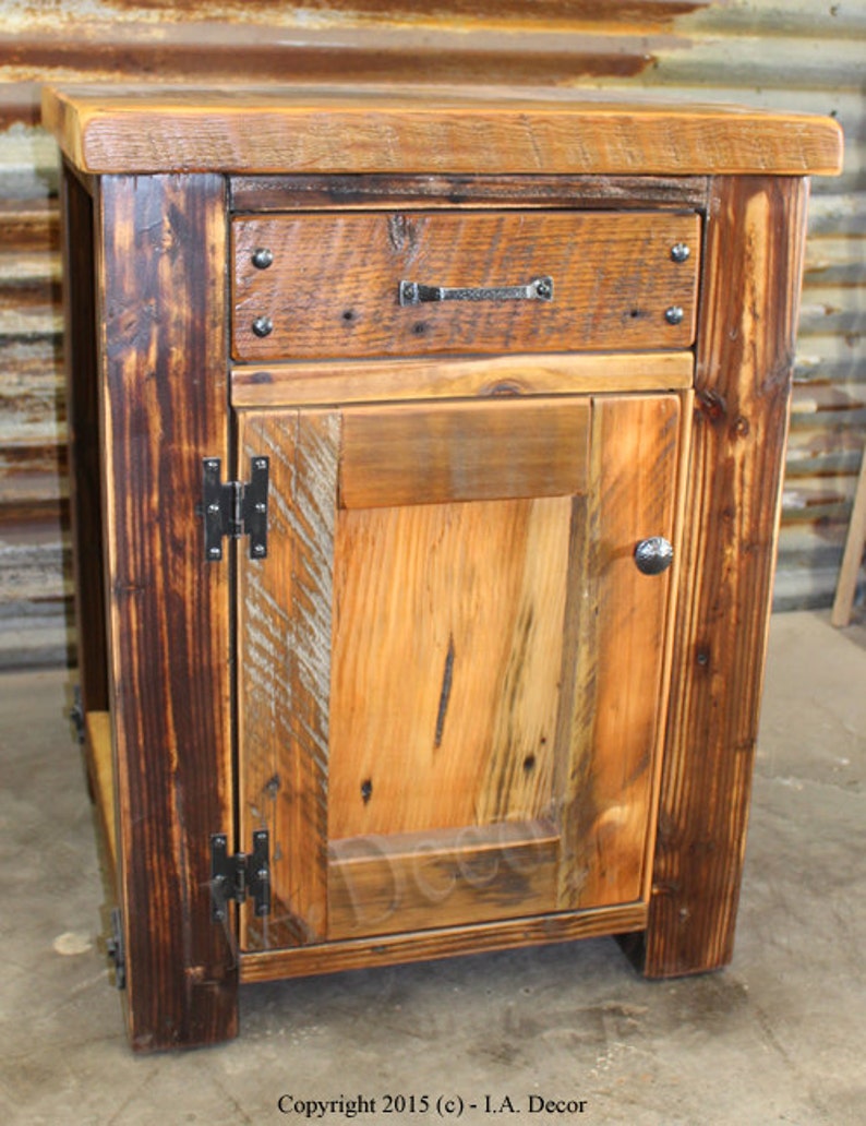 May include: A rustic wooden cabinet with a single drawer and a door. The cabinet is made of reclaimed wood and has a dark brown finish. The door has a simple design with a single panel and a black metal handle. The drawer has a black metal pull. The cabinet is sitting on a concrete floor.