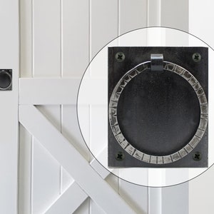 May include: Black metal door pull with a circular handle and a square backplate. The handle has a textured, hammered finish.