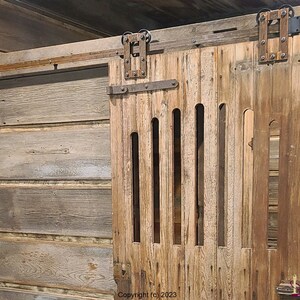 May include: A sliding barn door made of weathered brown wood featuring vertical slats and a series of narrow, elongated cutouts. The door is mounted on a dark metal track system with exposed hardware and rollers at the top.