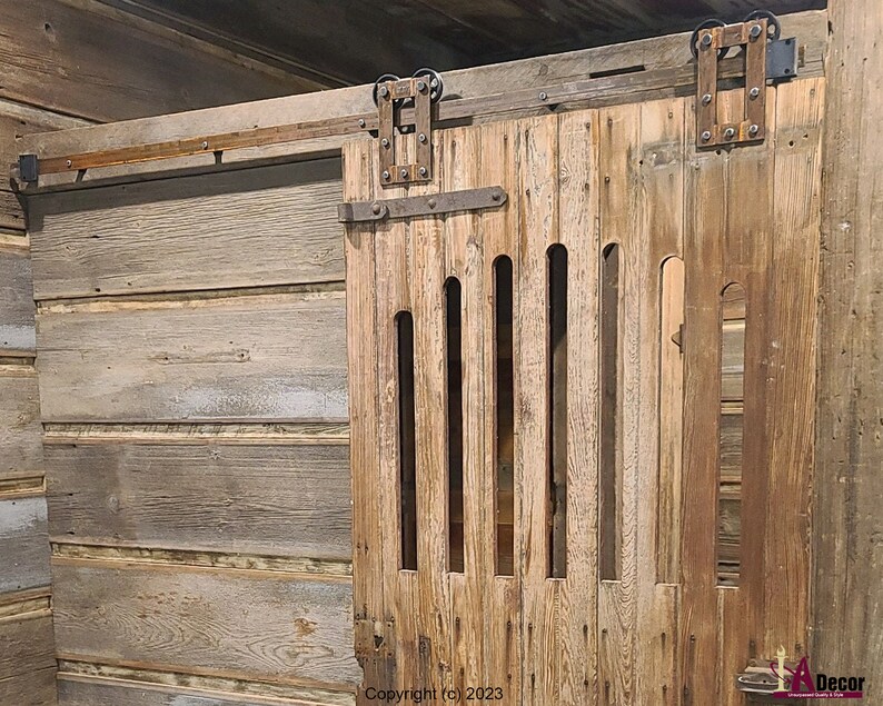 May include: A sliding barn door made of weathered brown wood featuring vertical slats and a series of narrow, elongated cutouts. The door is mounted on a dark metal track system with exposed hardware and rollers at the top.