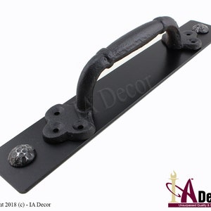 May include: Black iron door pull with a curved handle and decorative rosette accents. The pull is mounted on a rectangular plate. The handle is approximately 6 inches long.