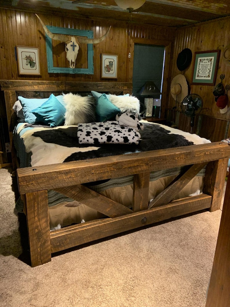 May include: A rustic wooden bed frame with a headboard and footboard. The bed features a cowhide blanket, teal cushions, and white fluffy cushions. The room has a western theme. The bed is made of dark brown wood.