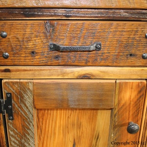 May include: Close-up of a wooden cabinet with a drawer and a door. The drawer has a black metal handle and the door has black metal hinges. The wood is a rustic brown color with a natural finish.