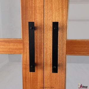 May include: Two black metal door handles are mounted on a wooden door. The handles are rectangular and have a simple design.