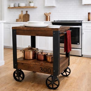 May include: A rectangular kitchen cart with a dark wood top and bottom shelf, supported by a black metal frame. The lower shelf features the number 300 printed in black. It has four black metal wheels and a red towel hanging from the side handle.