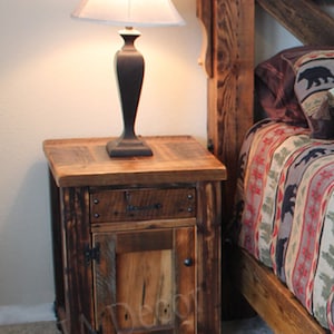 May include: A rustic wooden nightstand with a single drawer and a door. The nightstand is made of reclaimed wood and has a dark brown finish. It features a simple design with a single drawer and a door. The nightstand is sitting next to a bed with a rustic wooden headboard.
