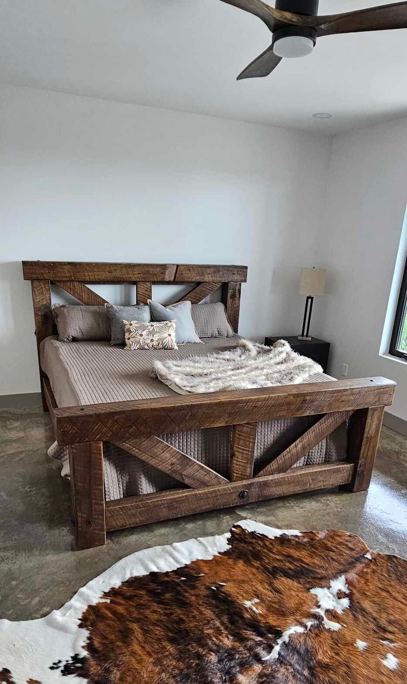 May include: A rustic wooden bed frame with a headboard and footboard, featuring a cross-beam design. The bed is made of dark brown wood and is covered with a brown comforter, pillows, and a faux fur throw. A cowhide rug is on the floor.