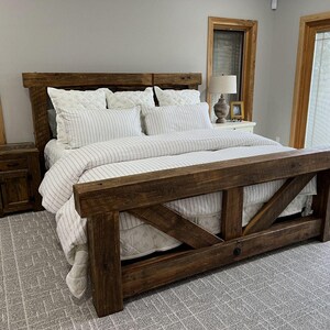 May include: A rustic wooden bed frame with a headboard and footboard. The bed is made of dark brown wood and features a mattress with white bedding and striped duvet cover. The room has a neutral colour scheme with a grey carpet and wooden accents.