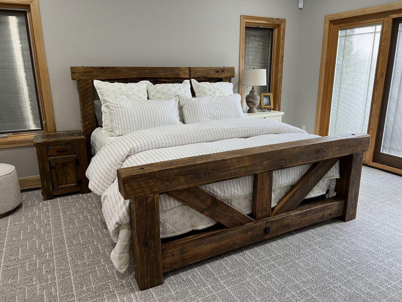 May include: A rustic wooden bed frame with a headboard and footboard. The bed is made of dark brown wood and features a mattress with white bedding and striped duvet cover. The room has a neutral colour scheme with a grey carpet and wooden accents.