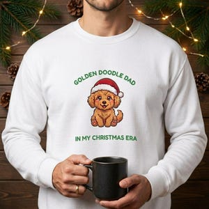 Może przedstawiać: Biała bluza z okrągłym dekoltem z grafiką brązowego szczeniaka w czerwono-białej czapce Mikołaja. Tekst nad szczeniakiem brzmi GOLDEN DOODLE DAD, a poniżej IN MY CHRISTMAS ERA, napisane zielonymi wielkimi literami.