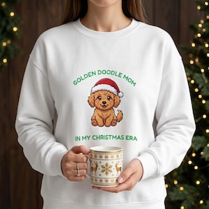 Może przedstawiać: Biała bluza z długim rękawem z grafiką szczeniaka golden doodle w czerwono-białej czapce Mikołaja. Napis GOLDEN DOODLE MOM IN MY CHRISTMAS ERA jest w kolorze zielonym. Trzymany jest biały kubek ze złotymi wzorami reniferów i płatków śniegu.