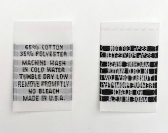 Op de afbeelding: Wit stoffen label met zwarte tekst. Het label vermeldt "65% Katoen 35% Polyester Machinewas koud Drogen in de droger op lage temperatuur Onmiddellijk verwijderen Niet bleken Gemaakt in de VS."