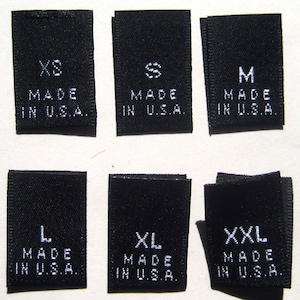 May include: Six black fabric labels with white text. The labels are for clothing sizes XS, S, M, L, XL, and XXL. Each label reads "Made in U.S.A."