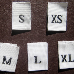 Könnte beinhalten: Sechs weiße Stoff-Etiketten mit schwarzer Beschriftung, die verschiedene Größen angeben: XS, S, M, L, XL.