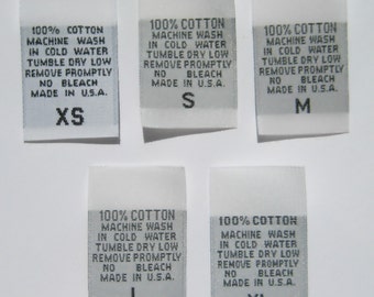 Peut inclure: Cinq étiquettes de taille de vêtements en tissu blanc avec du texte noir. Les étiquettes indiquent la taille, "XS", "S", "M", "L", et "XL". Chaque étiquette comprend les instructions : "100% Coton, Lavage en machine à l'eau froide, Séchage en machine à basse température, Retirer rapidement, Pas de blanchiment, Fabriqué aux États-Unis."
