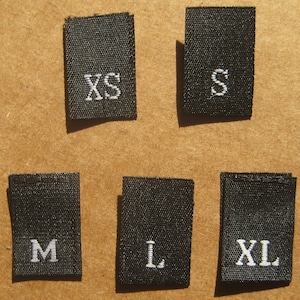 Könnte beinhalten: Fünf schwarze Stoff-Etiketten mit weißer Beschriftung, die die Größen XS, S, M, L und XL angeben.