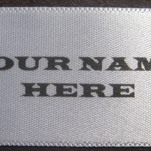 Puede incluir: Una etiqueta de sat&eacute;n blanco con texto negro que dice "YOUR NAME HERE".
