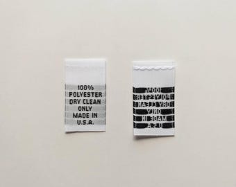 Peut inclure: Deux étiquettes en tissu blanc avec du texte noir. La première étiquette indique "100% Polyester Nettoyage à sec uniquement Fabriqué aux États-Unis". La deuxième étiquette comporte une série de lignes noires avec des chiffres et des lettres.
