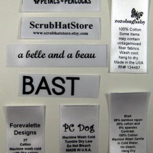 May include: A collection of white satin labels with black text. The labels include the names of different businesses, such as "Forevalette Designs", "PC Dog", and "ScrubHatStore". Some labels include care instructions, such as "Machine Wash Cold" and "Tumble Dry Low".