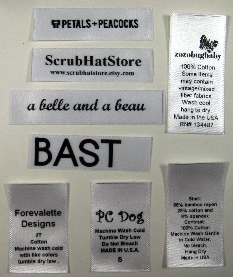 May include: A collection of white satin labels with black text. The labels include the names of different businesses, such as "Forevalette Designs", "PC Dog", and "ScrubHatStore". Some labels include care instructions, such as "Machine Wash Cold" and "Tumble Dry Low".