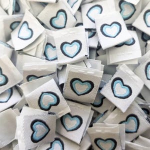 May include: A pile of white fabric labels with a blue and white heart design. The labels are square shaped and have a black outline around the heart.
