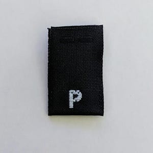 May include: A small, rectangular black fabric label with a white embroidered "P" in the lower center. The label has a woven texture and appears to be a clothing tag or patch.