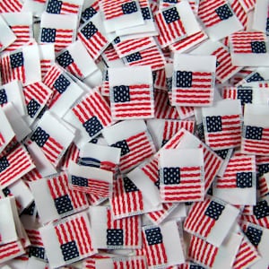 May include: A pile of small white fabric squares with an embroidered American flag design in red, white, and blue.