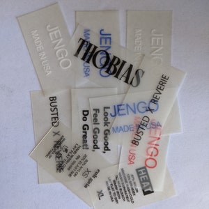 May include: A stack of white, semi-transparent clothing labels with black text. The labels are for the brand "JenGo" and include the text "Made in USA", "Busted X Reverie", "Heat", "Look Good, Feel Good, Do Great!" and "Cult Gaia".