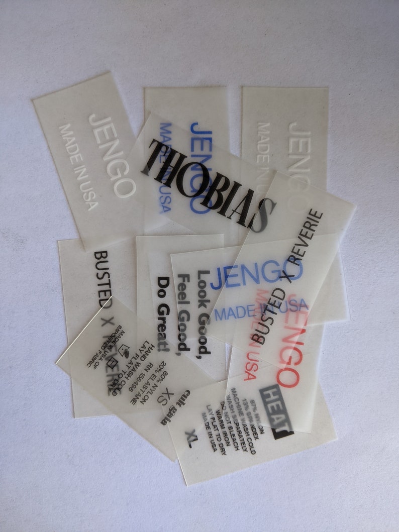 May include: A stack of white, semi-transparent clothing labels with black text. The labels are for the brand "JenGo" and include the text "Made in USA", "Busted X Reverie", "Heat", "Look Good, Feel Good, Do Great!" and "Cult Gaia".