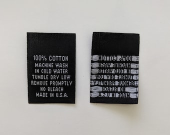 Op de afbeelding: Twee zwarte stoffen labels met witte tekst. Het label aan de linkerkant heeft de tekst "100% Katoen, Machinewas koud, Droogtrommel lage stand, Snel verwijderen, Geen bleekmiddel, Gemaakt in de VS". Het label aan de rechterkant heeft een reeks witte lijnen.