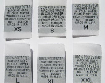 May include: Six white fabric clothing size tags with black text. The tags are labeled with sizes XS, S, M, L, XL, and XXL. Each tag includes the text "100% Polyester, Machine Wash in Cold Water, Tumble Dry Low, Remove Promptly, No Bleach, Made in U.S.A."