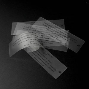 May include: Close-up of several transparent clothing care labels with black text. The labels contain washing instructions and the text "Work in Process" and "Made in USA with Imported Fabrics".