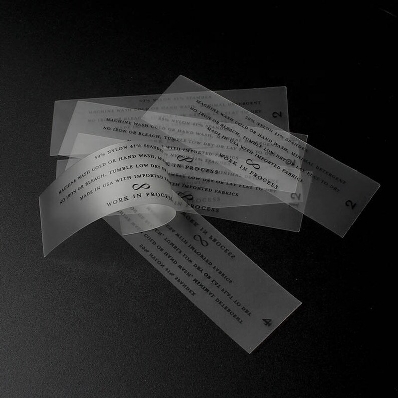 May include: Close-up of several transparent clothing care labels with black text. The labels contain washing instructions and the text "Work in Process" and "Made in USA with Imported Fabrics".