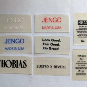 May include: Eight white rectangular clothing labels with black text. The labels include the words "JENGO Made in USA", "Look Good, Feel Good, Do Great!", "BUSTED X REVERIE", "THOBIAS", "HEAT 87% Nylon 13% Spandex Machine Wash Cold Wash Separately Do Not Bleach Warm Iron Lay Flat to Dry Made in USA XL", and "cuk gala XS 80% Nylon 20% Elastane RN 158496 Hand Wash Cold Lay Flat to Dry Made in USA of Imported Fabric".