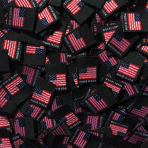 May include: A close-up of a pile of black fabric labels with the text "Made in USA" printed in white. The labels are arranged in a random pattern and some are overlapping.