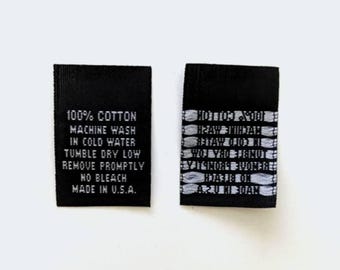 Op de afbeelding: Twee zwarte stoffen labels met witte tekst. Het label aan de linkerkant luidt "100% Katoen, Machinewas koud, Droogtrommel lage stand, Snel verwijderen, Niet bleken, Gemaakt in de VS". Het label aan de rechterkant heeft een herhalend patroon van witte tekst.